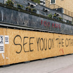 En person cyklar förbi ett högt träplank med där det står skrivet med svart See you on the other side