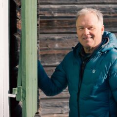 En medelålders man i blå täckjacka står vid en husvägg med träpanel och håller i en grön fönsterlucka.