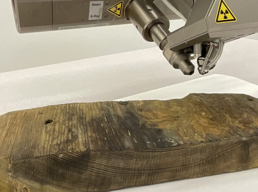 En metallfärgad röntgenapparat undersöker en bit av en mörkfärgad tjock planka.