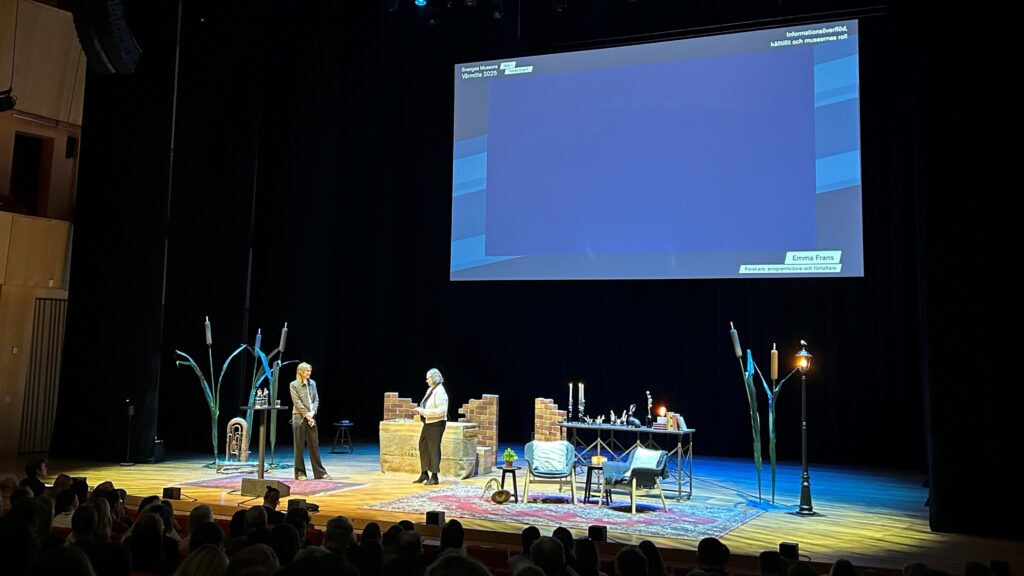 Två personer på en scen. Långt avstånd. Ovanför dem båda en blålysande projektorskärm.