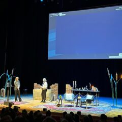 Två personer på en scen. Långt avstånd. Ovanför dem båda en blålysande projektorskärm.