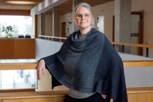 Kvinna med grått hår och grå poncho står lutad mot en balustrad i en trapphall.