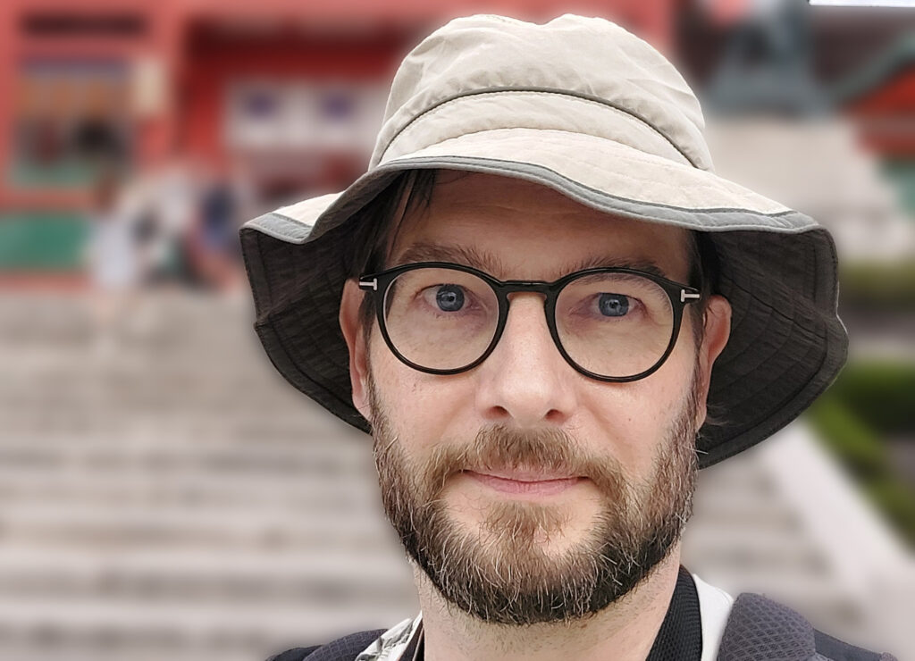 En man med glasögon i Khakifärgad hatt tittar in i kameran. I bakgrunden en bred trappa upp mot en byggnad, som man bara ser grunden på.