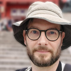 En man med glasögon i Khakifärgad hatt tittar in i kameran. I bakgrunden en bred trappa upp mot en byggnad, som man bara ser grunden på.