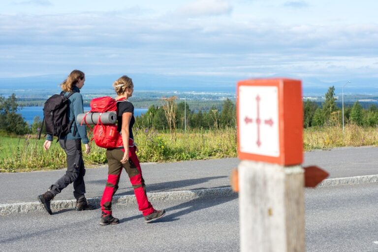 Två personer med vandringsskor och ryggsäckar går på S:t Olavsleden som är utmärkt med en stolpe med ett kors av pilar på.