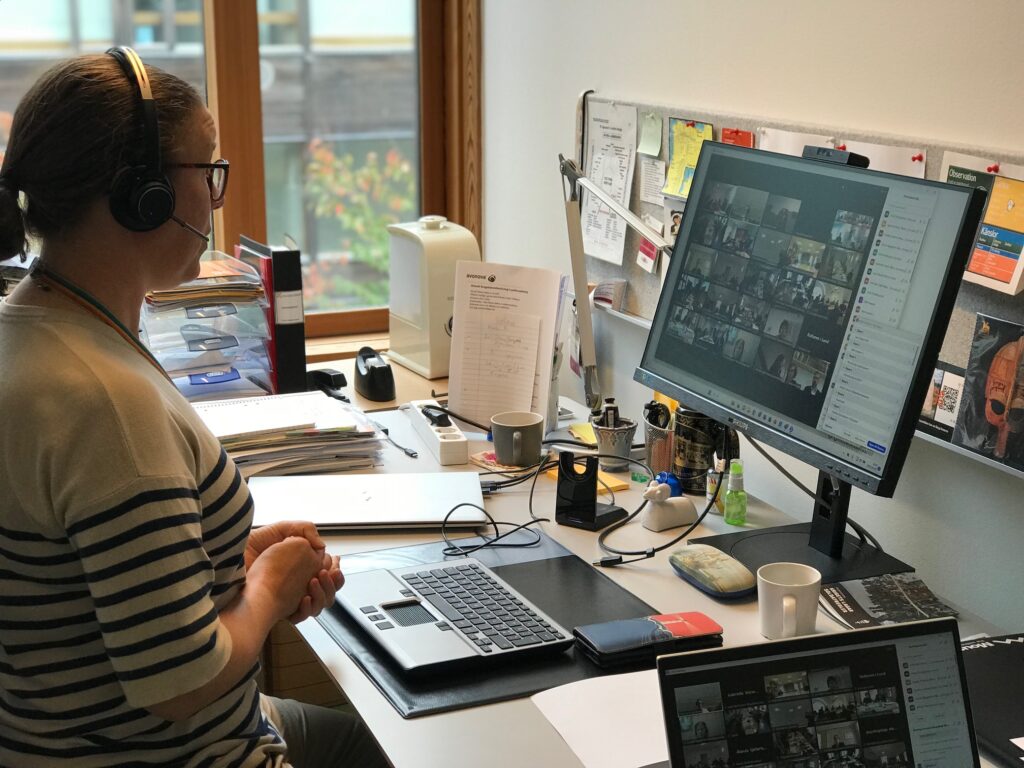 En kvinna sitter framför ett möte vid datorn. På skärmen skymtar många deltagare.
