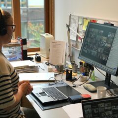 En kvinna sitter framför ett möte vid datorn. På skärmen skymtar många deltagare.