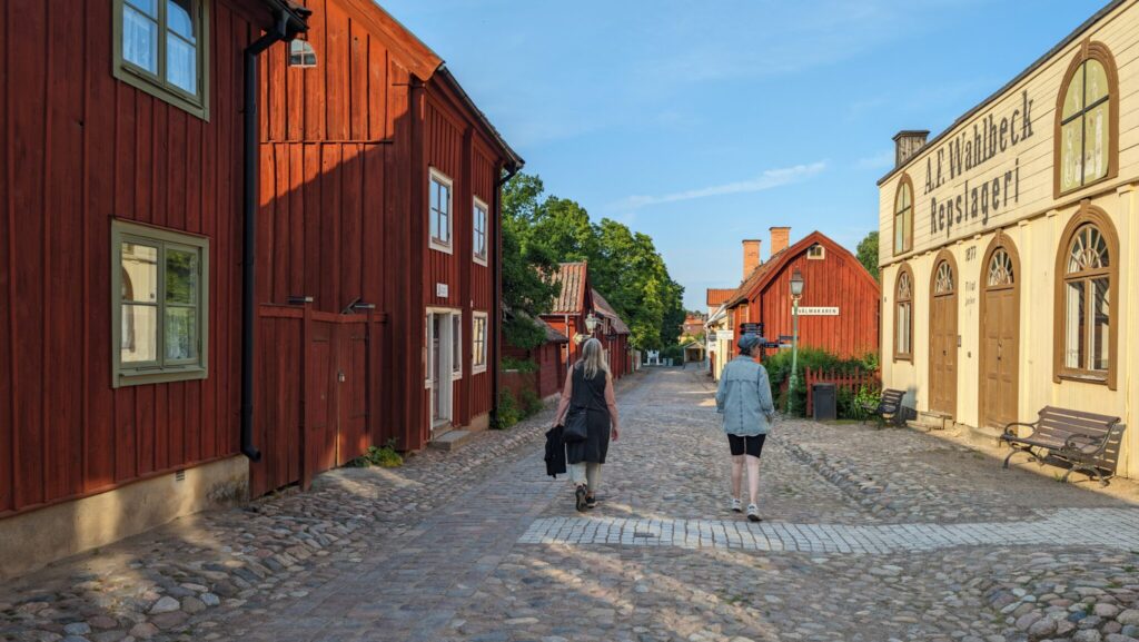 Två damer vandrar i gamla kvarter på en bred kullerstensgata omgiven av äldre träbyggnader.