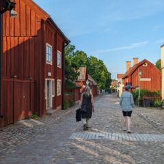 Två damer vandrar i gamla kvarter på en bred kullerstensgata omgiven av äldre träbyggnader.