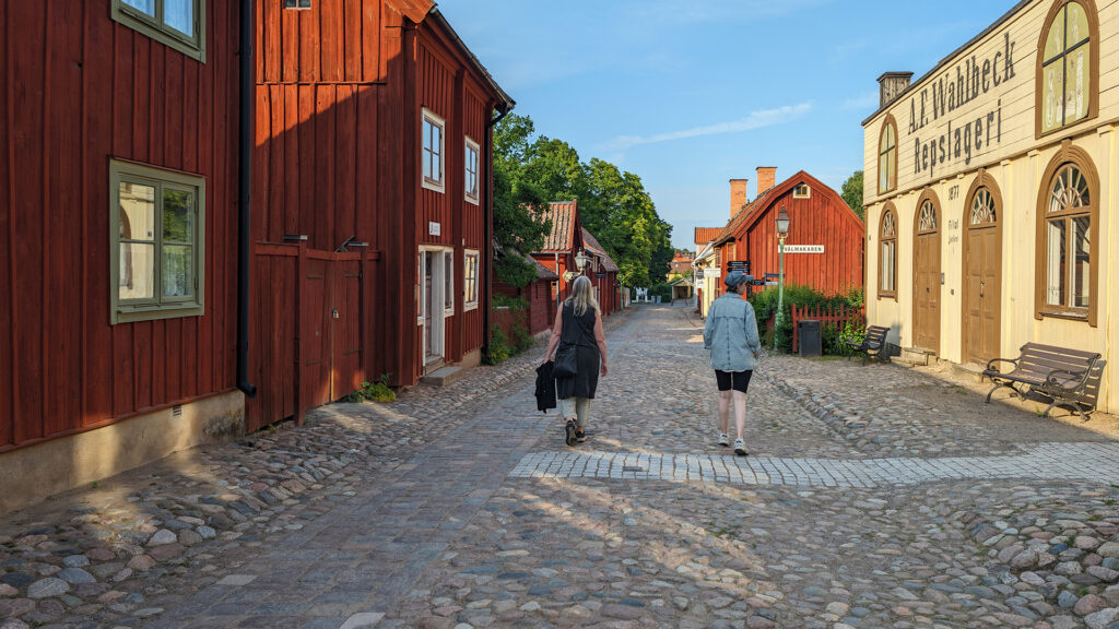 Två damer vandrar i gamla kvarter på en bred kullerstensgata omgiven av äldre träbyggnader.