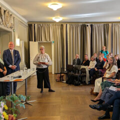 En seminariesal med draperier i bakgrunden. Till vänster står en panel och diskuterar. Till höger sitter åhörare och lyssnar. En av dem är Kungen.