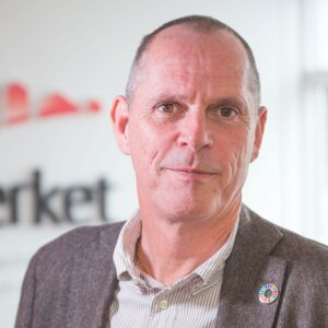 Porträtt på man, kort grått hår, brun kavaj och skjorta, inne.