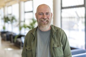 Porträtt på en man, grått kort hår och skägg, grå tröja, grön jacka. Inne, ljus korridor på ett kontor med stora fönster.