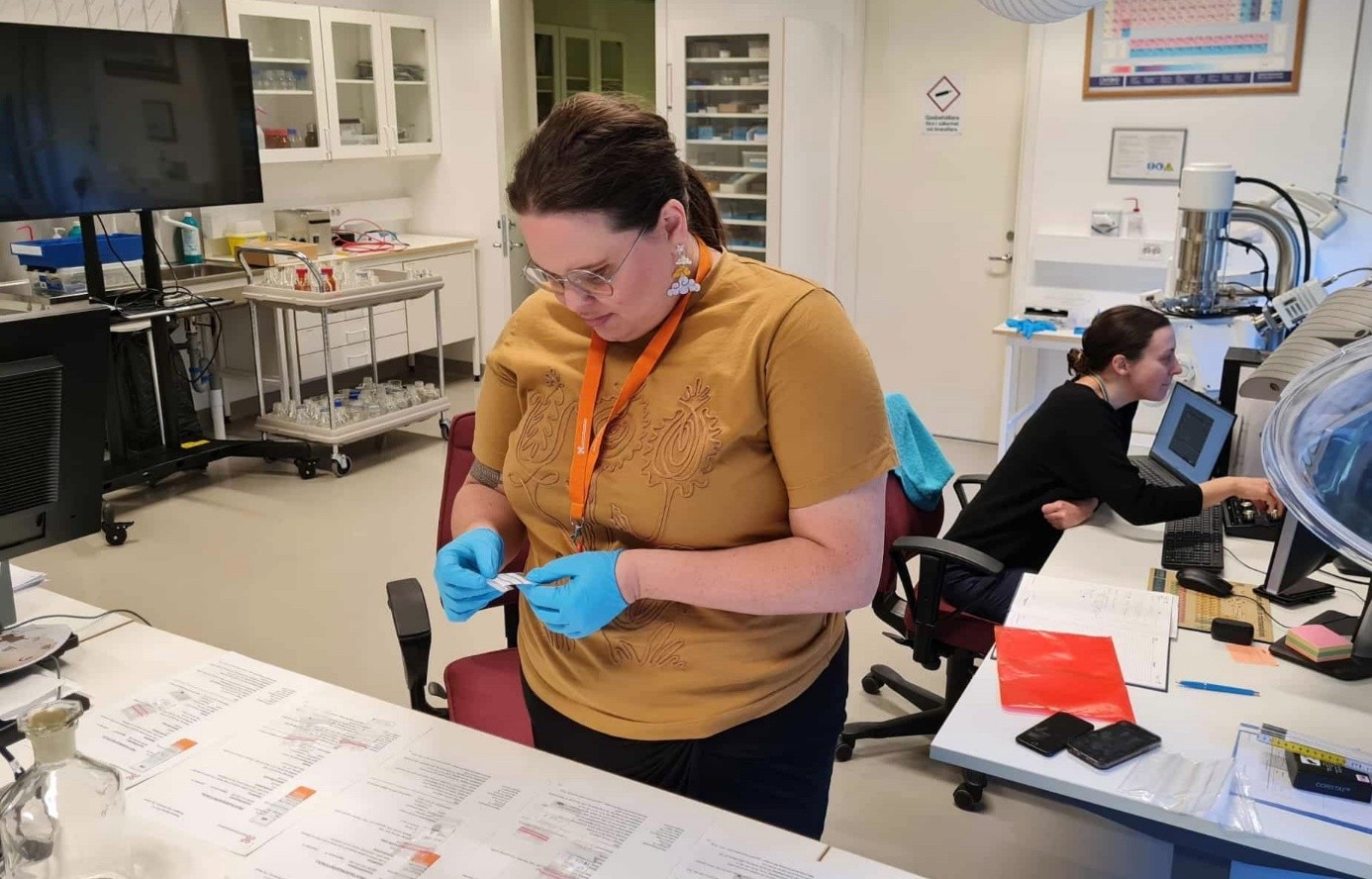 Laboratoriemiljö med två personer i arbete. En person i förgrunden bär blå handskar och undersöker ett objekt, medan en annan person i bakgrunden sitter vid en dator. Rummet innehåller laboratorieutrustning, hyllor med material och dokument utspridda på borden.