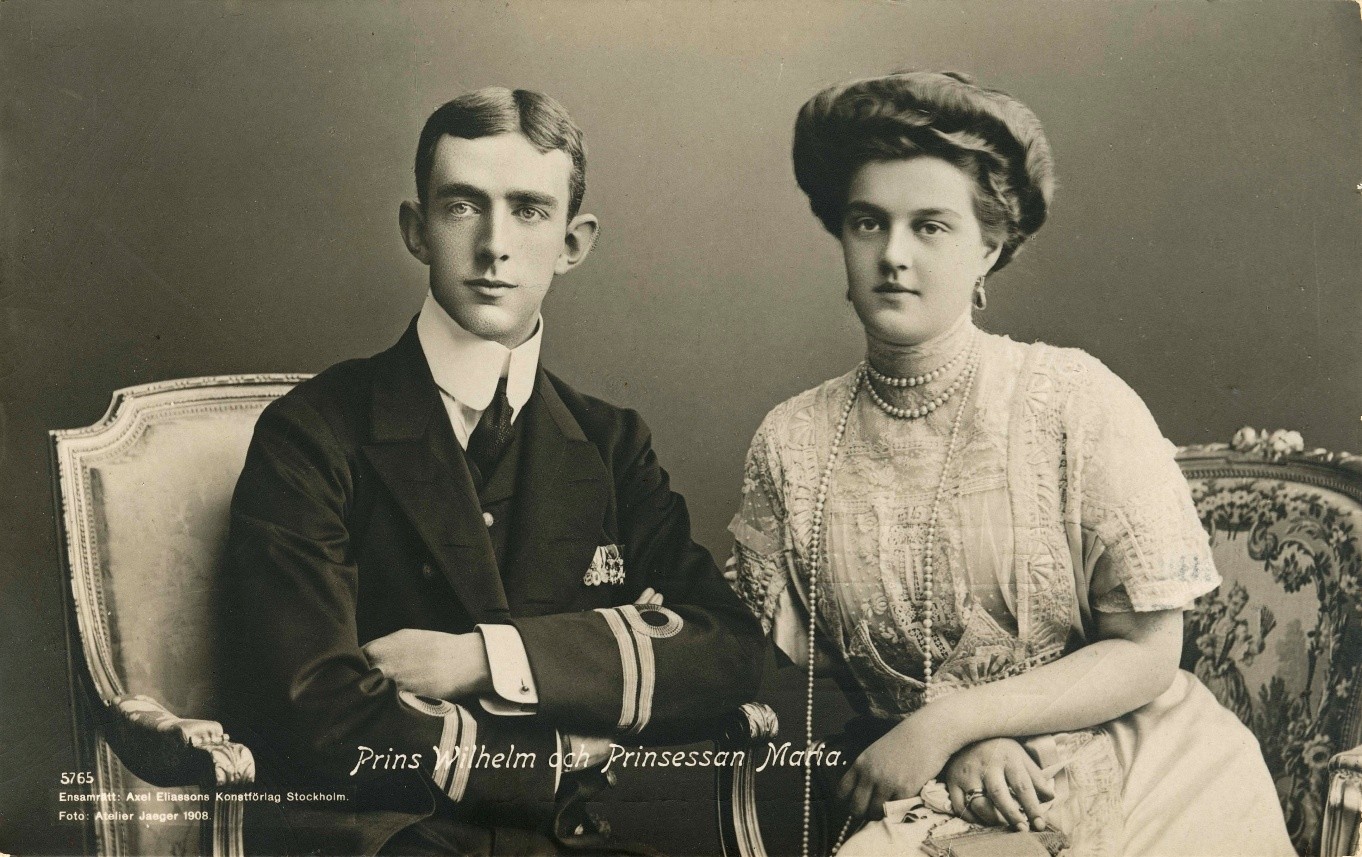Två personer sitter på utsmyckade stolar. Den vänstra personen bär mörk kostym med vit skjorta och slips, med armarna i kors. Den högra personen bär en ljus klänning med intrikata mönster och flera pärlhalsband. Ansiktena är skymda. Texten i bilden lyder: 'Prins Wilhelm och Prinsessan Maria. 5765 Ernst & Alice Eklund, Konserthog Stockholm Foto Atelier Jaeger 1908.