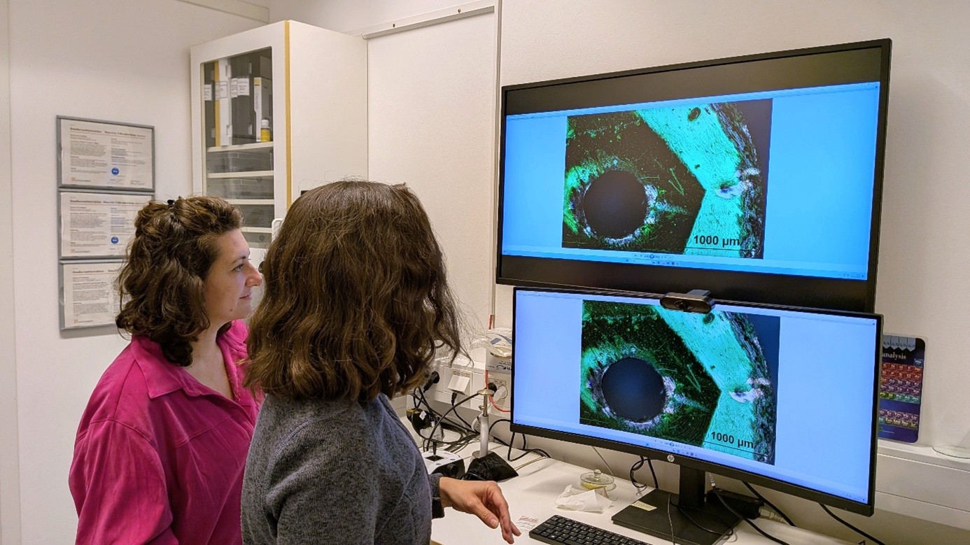 Två personer står framför ett skrivbord med två datorskärmar som visar samma mikroskopbild. Bilden visar ett material med ett cirkulärt hål och grönaktig färgning, samt en skalmarkering som anger '1000 µm'. Rummet är ett kontor eller laboratorium med utrustning och dokument i bakgrunden.