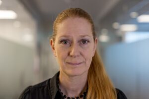 Porträttbild på kvinna med rött hår uppsatt i hästsvans och mörk kavaj. Inne.