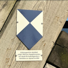 En blåvit skylt. Under skylten står texten "Kulturegendom skyddad enligt 1954 års Haagkonvention..."