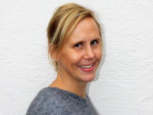 porträtt kvinna, mellanblont halvlångt hår, grå tröja, inne, vit vägg i bakgrunden.