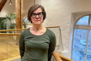 Porträtt kvinna, halvkort brunt hår, glasögon och grön klänning. Inne, stående i ett kontorslandskap.