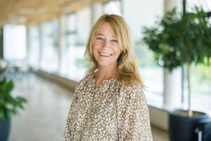 Porträtt, kvinna med ljust halvlångt hår och mönstrad blus. inne i en korridor med stora fönster i kontorsmiljö.