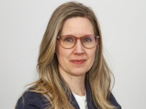 porträtt, kvinna med glasögon, långt mellanblont hår och mörk kavaj. Grå bakgrund.