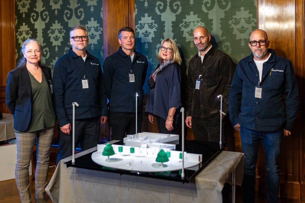 Sex personer står bakom ett bord med en arkitektonisk modell som visar ett utomhusområde med träd och strukturer, troligen ett parkområde eller stadsutvecklingsprojekt. Personernas ansikten är suddade för att skydda deras integritet. Bakgrunden består av träpaneler och mönstrad tapet