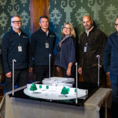 Sex personer står bakom ett bord med en arkitektonisk modell som visar ett utomhusområde med träd och strukturer, troligen ett parkområde eller stadsutvecklingsprojekt. Personernas ansikten är suddade för att skydda deras integritet. Bakgrunden består av träpaneler och mönstrad tapet