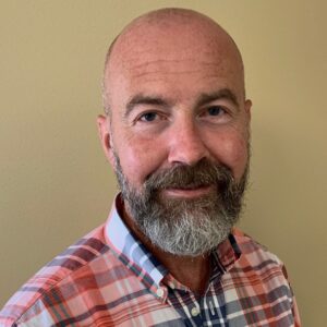 Porträtt, man med grånande skägg och rutig skjorta, inne, gul vägg i bakgrunden.