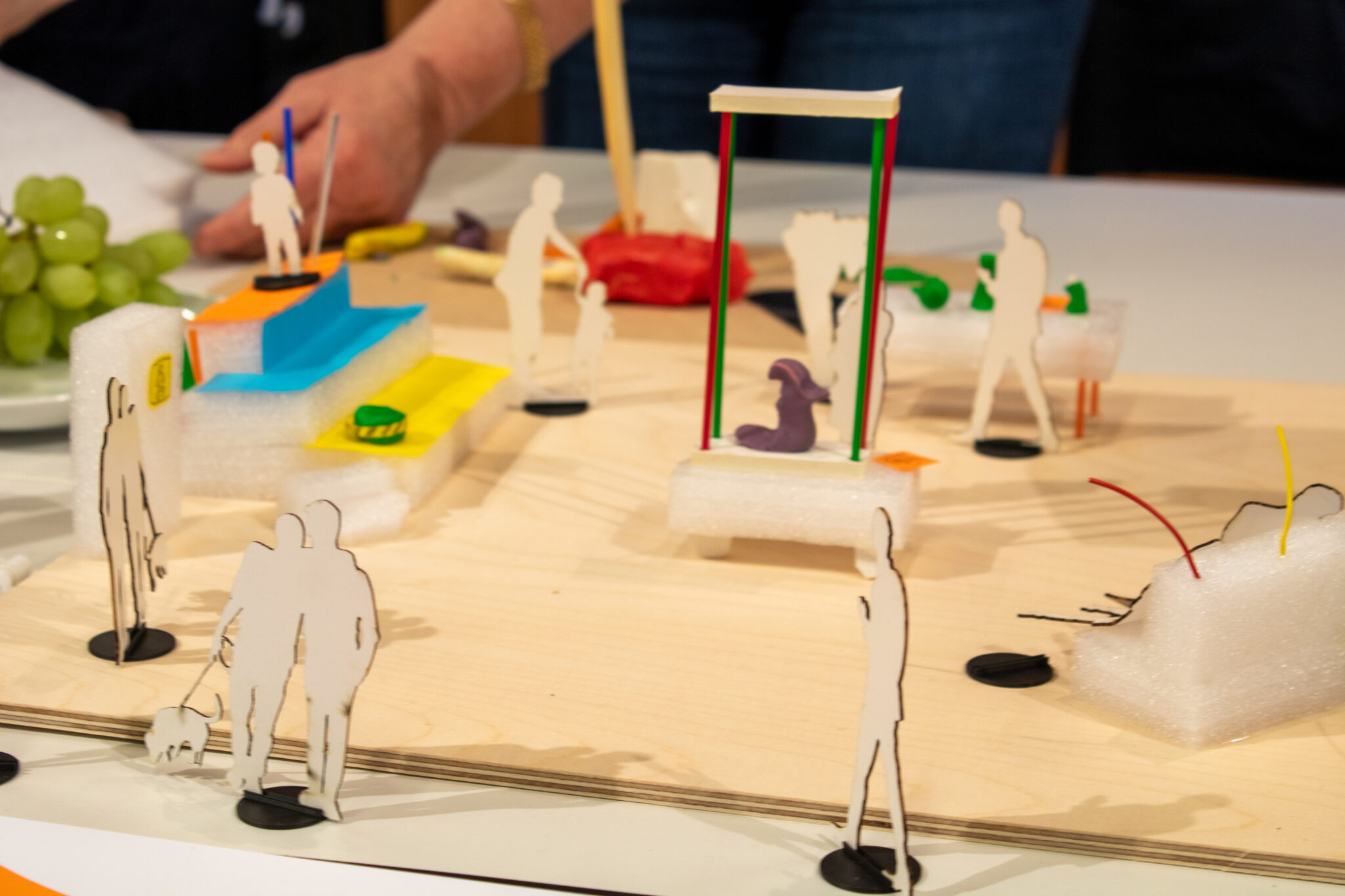 Miniatyrmodell av en arkitektonisk miljö med små figurer av människor som står eller går. Modellen innehåller trappor, plattformar, ett gungställ med färgade stolpar, bänkar och abstrakta former. I bakgrunden syns en tallrik med gröna vindruvor och händer som arbetar med modellen.