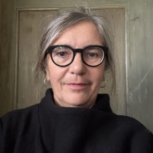 Porträtt av kvinna, grått uppsatt hår, svarta glasögonbågar och svart stickad tröja. inne, en målad dörr i bakgrunden.