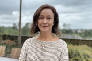 Porträtt, kvinna, axellångt brunt hår, ljsu tröja. Ute, lantlig miljö, himmel med moln.