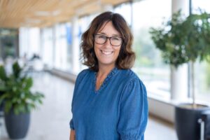 Porträtt av kvinna, mörkt halvlångt hår, glasögon, blå skjorta. Inne, en ljus korridor med stora fönster i bakgrunden.