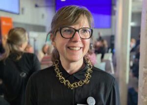 Porträtt kvinna, glasögon, svart blus, uppsatt hår. Inne. Folkvimmel i bakgrunden.