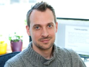 Porträtt man, kort mörkt hår, grå stickad polotröja, kontorsmiljö, inne.