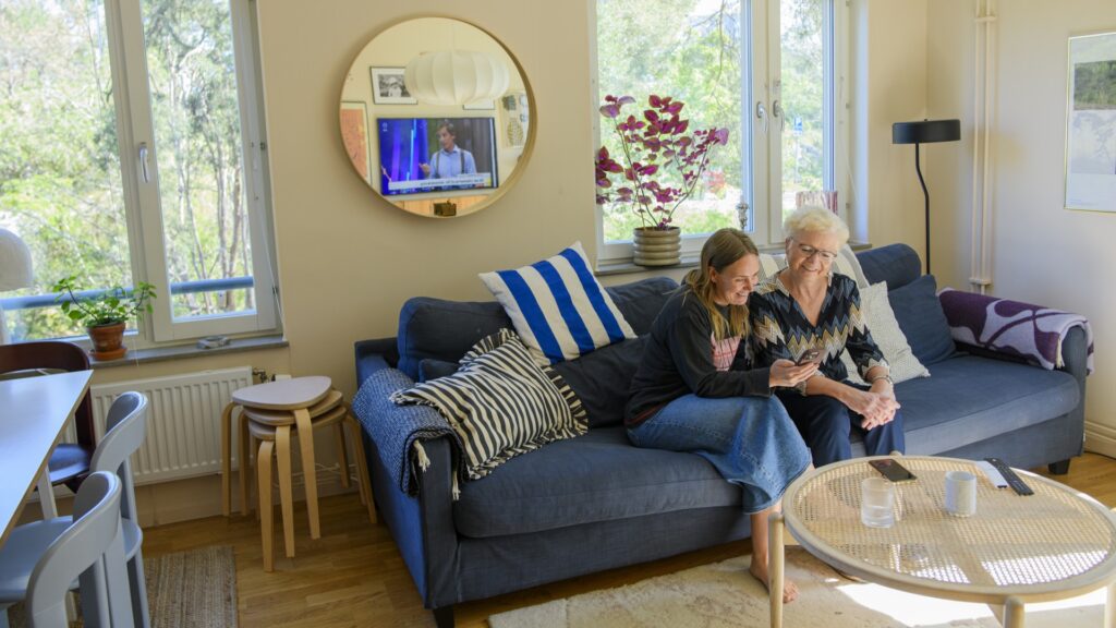 två personer sitter i en soffa i ett vardagsrum och tittar tillsammans på en mobiltelefon.