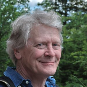 Porträtt, man grått hår, blå tröja, Ute, träd och buskar i bakgrunden.