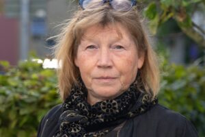 Porträtt av kvinna med axellång, brunt hår, svart schal om halsen och svart kappa, ute, grönska och byggnader i bakgrunden.