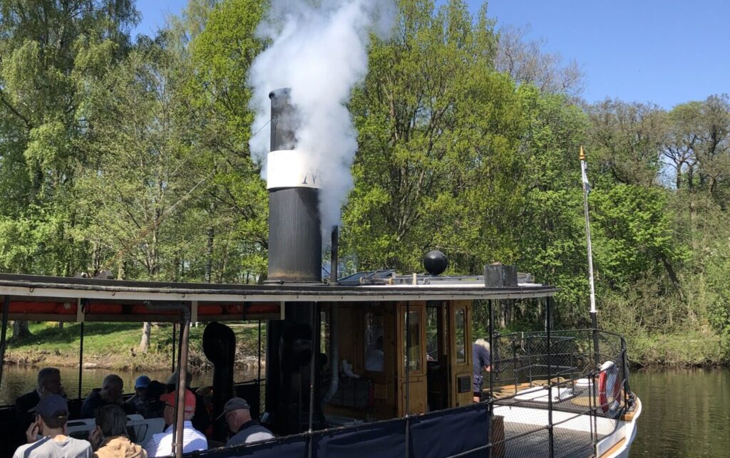 Delar av en ångbåt med öppna sidor ´där det sitter flera passagerare och tittar ut. På taket väller vit rök ut en skorsten .
