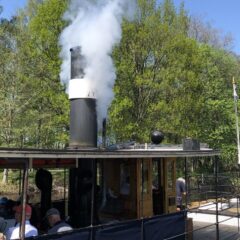 Delar av en ångbåt med öppna sidor ´där det sitter flera passagerare och tittar ut. På taket väller vit rök ut en skorsten .