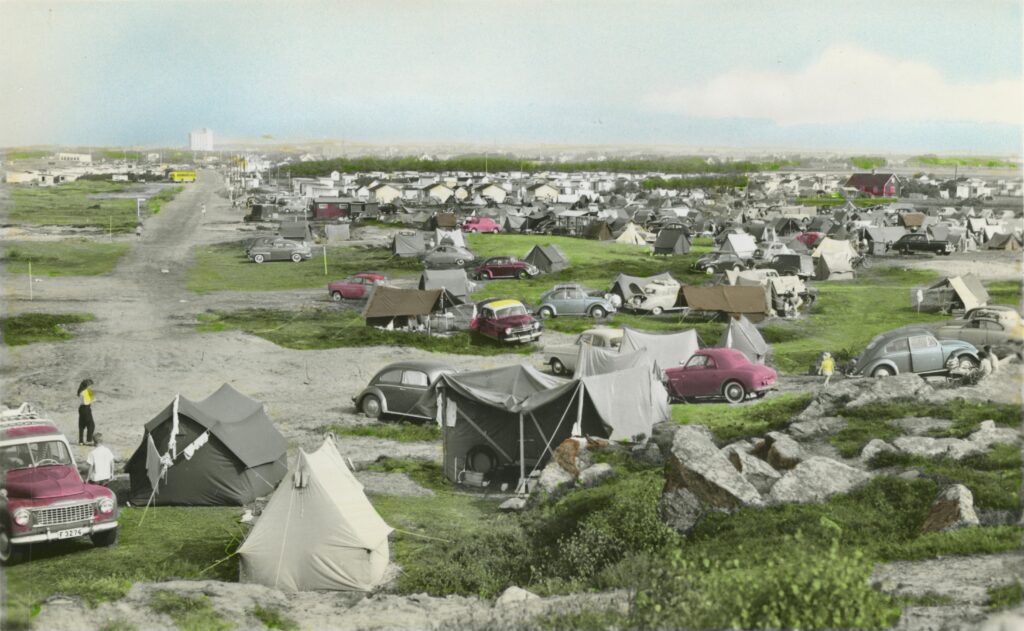 Tält och bilar står uppställda på en gräsbevuxen yta. I bakgrunden syns bebyggelse och vid horisonten reser sig en hög industribyggnad.