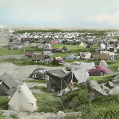 Tält och bilar står uppställda på en gräsbevuxen yta. I bakgrunden syns bebyggelse och vid horisonten reser sig en hög industribyggnad.