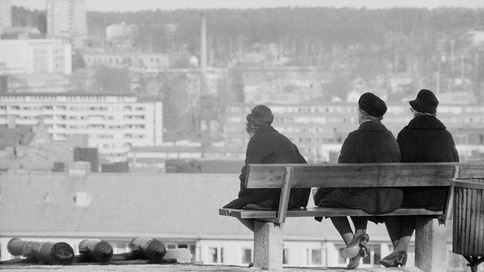 Svartvit bild med vy över ett hamninlopp och tre svartkädda damer med ryggen emot som sitter på en bänk och ser ut över vattnet.