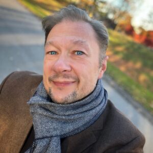 porträtt, man, mörkt hår, brun kavaj. uteväg och lantlig miljö med hus och höstlöv i bakgrunden.