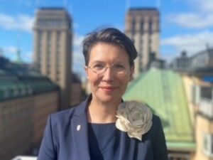 porträtt, kvinna, kort mörkt hår, glasögon, mörk kavaj med en ros på kavajslaget, ute, sol, byggnader i bakrunden.