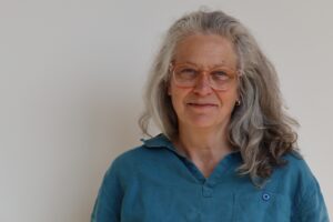 Porträtt av kvinna, glasögon, långt grått hår, blå skjorta. Inne. Vit vägg i bakgrunden.