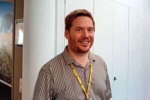 Porträtt av man, kort brunt hår, rutig skjorta, inne, vita skåp i bakgrunden i ett kontorslandskap.