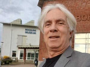 Selfiebild på en man med vitt hår och kavaj, ute, byggnader i bakgrunden.