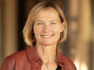 Porträtt kvinna, axellångt blont hår, röd skinnjacka, diffus bakgrund.