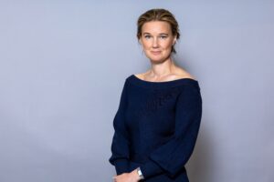 Porträtt kvinna i mörk dräkt, med brunt uppsatt hår och mörkblå dräkt. Grå bakgrund.